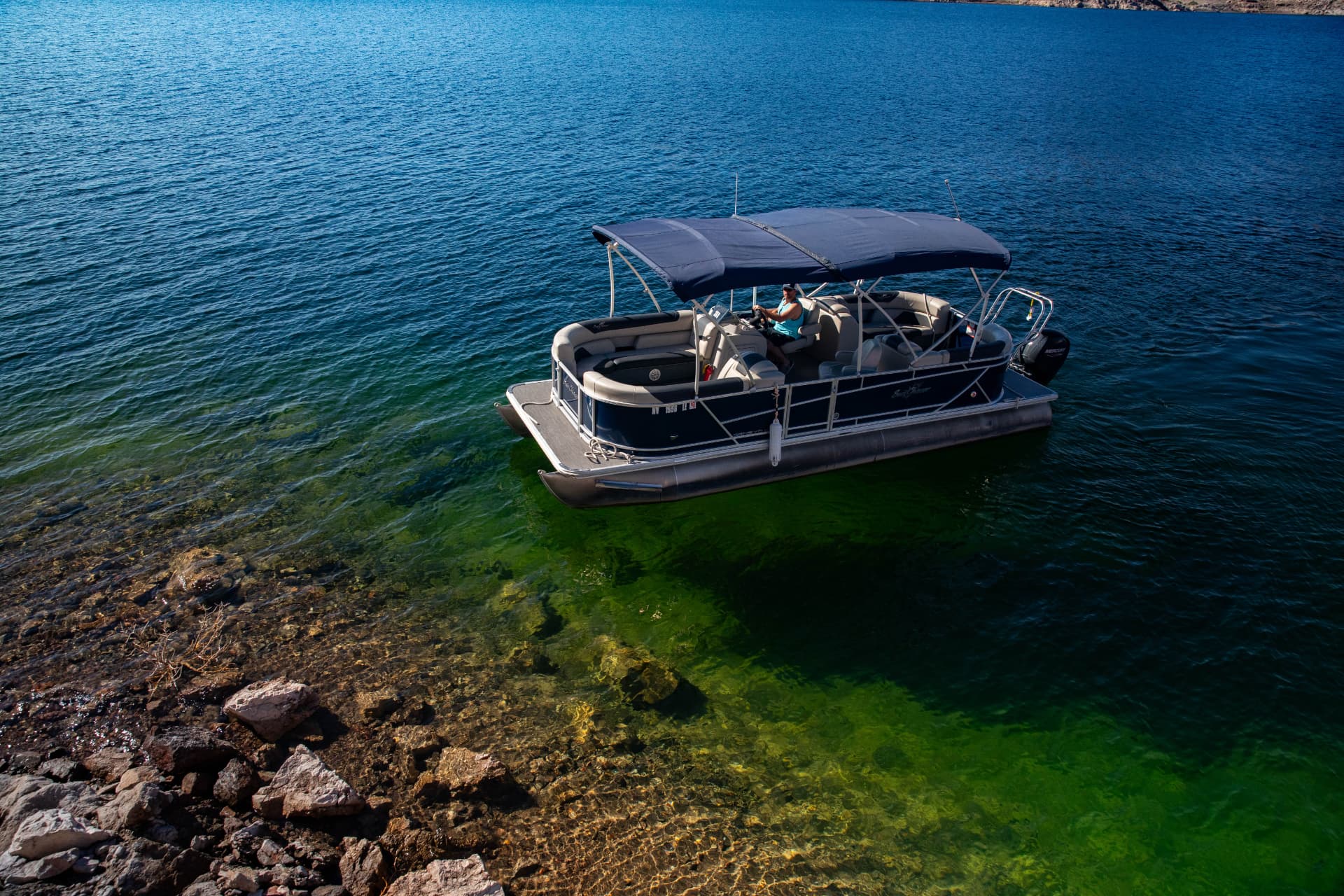 Navy pontoon boat with a shade canopy floating on clear green water beside a rocky Lake Mead shoreline